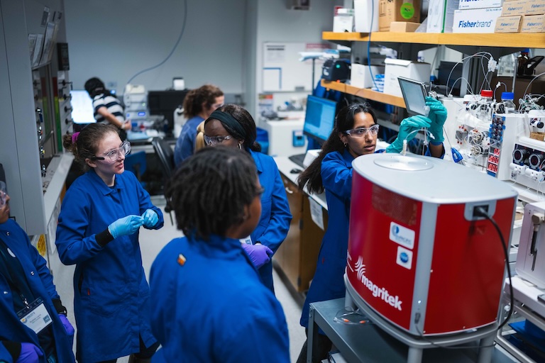A group of people wearing blue lab coats and gloves are working in a laboratory. They are gathered around various pieces of equipment, including a red machine labeled "Magritek." The lab is filled with shelves containing various supplies and equipment, and there are computer monitors on the desks. The faces of the individuals have been blurred for privacy.