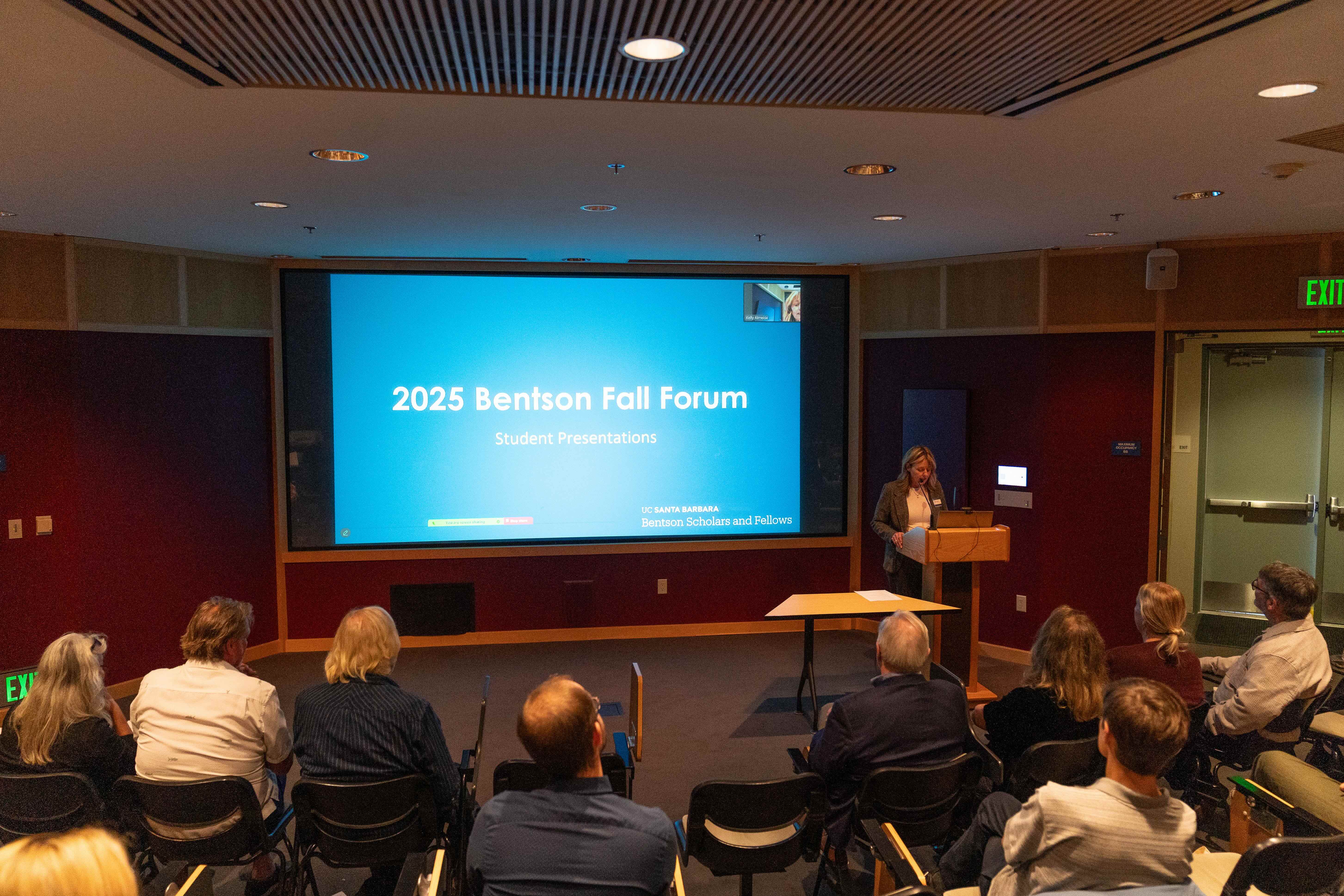 A speaker stands at a wooden podium addressing a seated audience in a conference room. A large projection screen displays a blue slide with white text reading "2025 Bentson Fall Forum: Student Presentations" alongside the UC Santa Barbara logo.