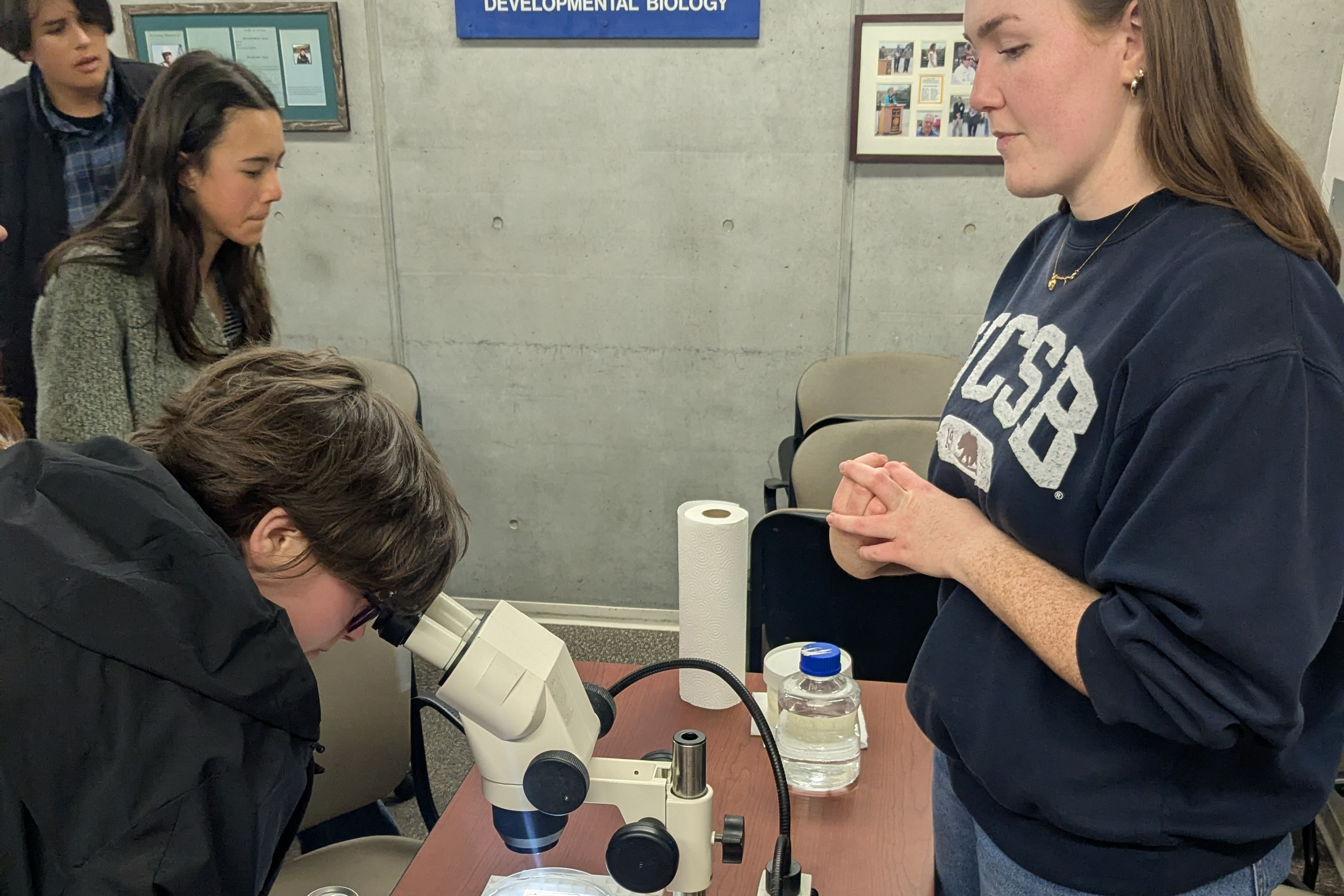 A UCSB mentor in a UCSB sweatshirt speaks with a student looking through a microscope.