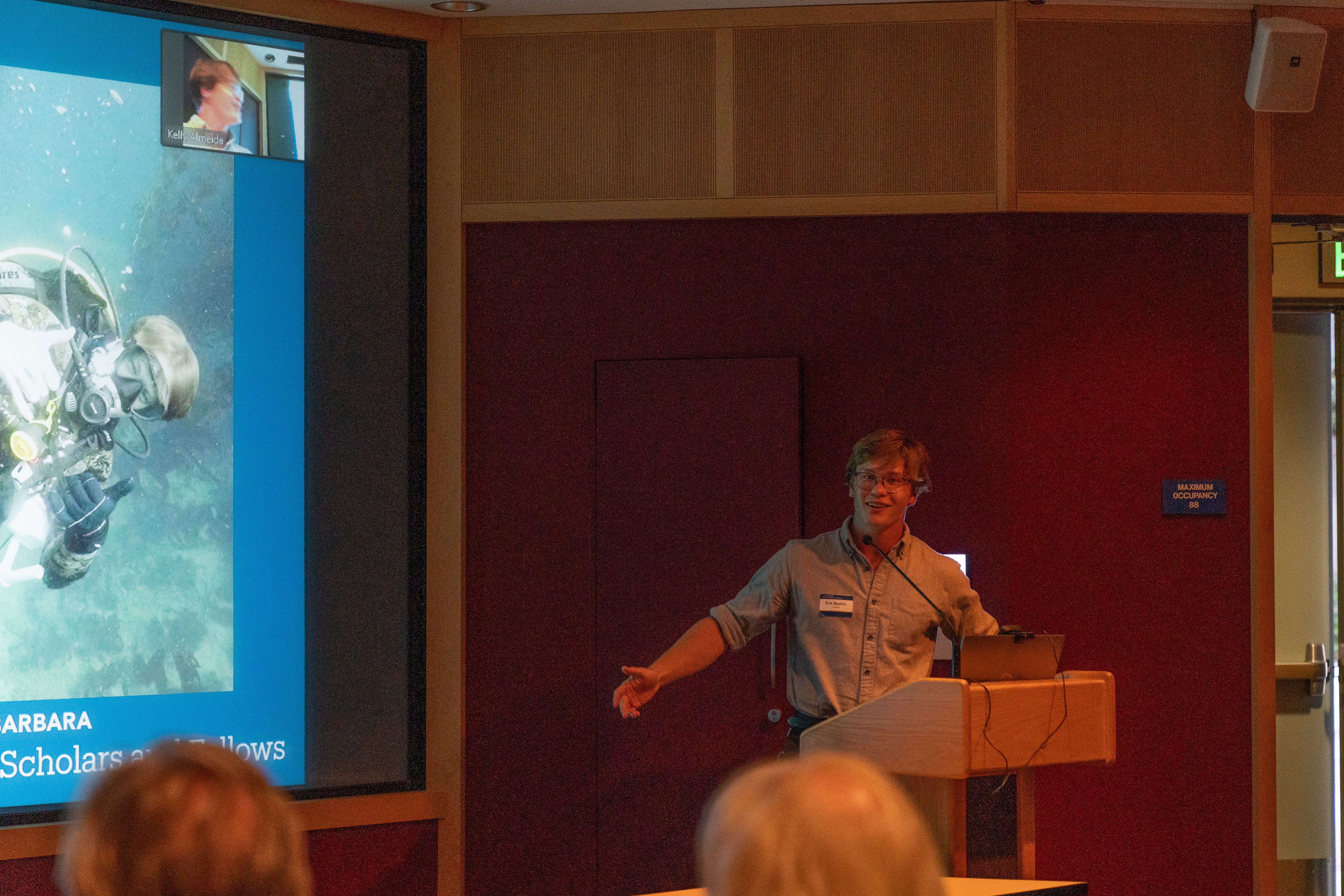 A smiling student stands at a podium, gesturing with his left hand during a presentation. The screen behind him displays a large photo of a scuba diver underwater, along with a smaller video feed of a remote participant in the upper corner.