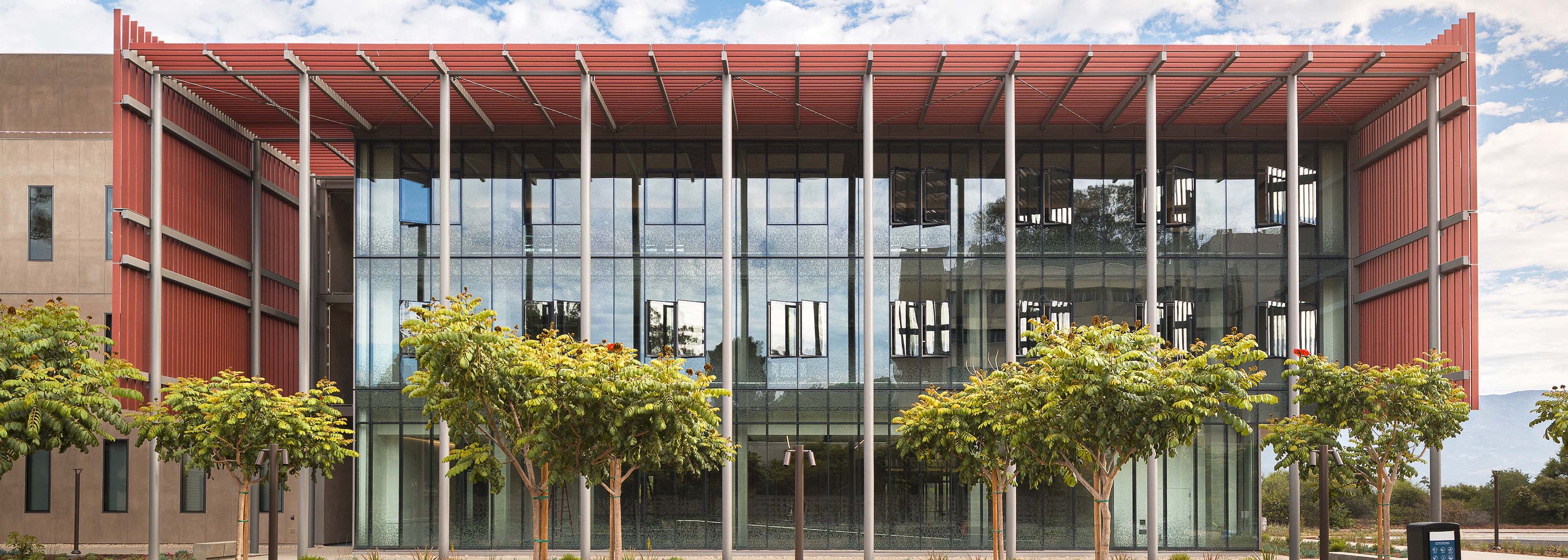 Exterior of Henley Hall at UC Santa Barbara featuring large glass windows, a reddish-brown slatted roof overhang, and small trees in the foreground.