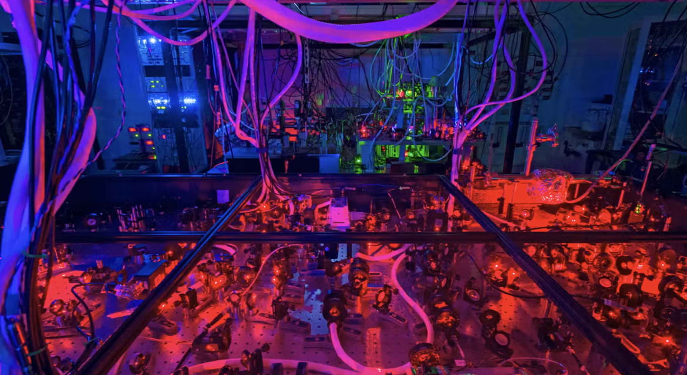 A dense optical table setup in David Weld's physics laboratory, illuminated by glowing red and purple lights, featuring mirrors and lenses.