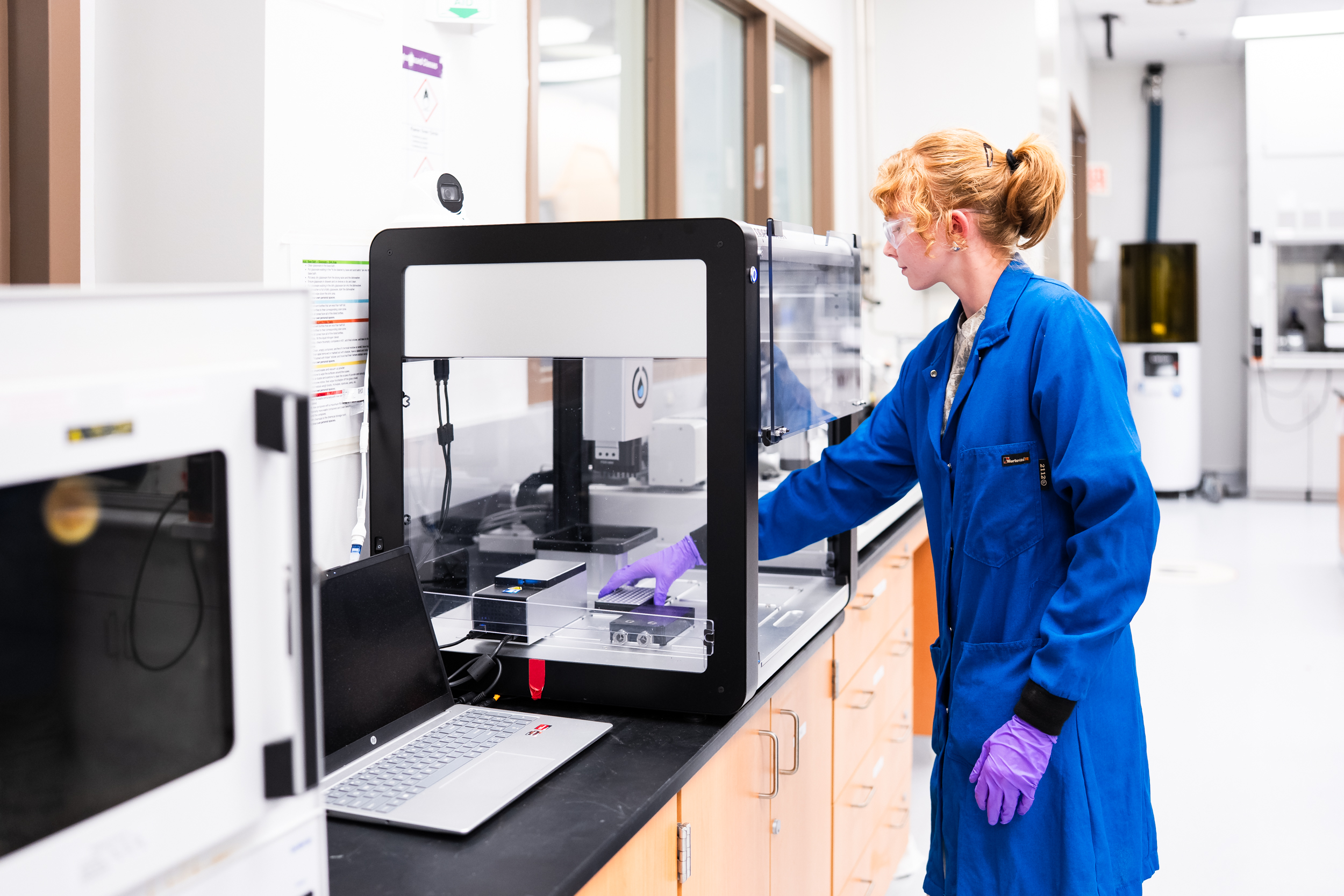 Lab assistant Marissa Beyer, wearing safety glasses, a blue lab coat, and purple gloves, loads a robotic liquid handler.