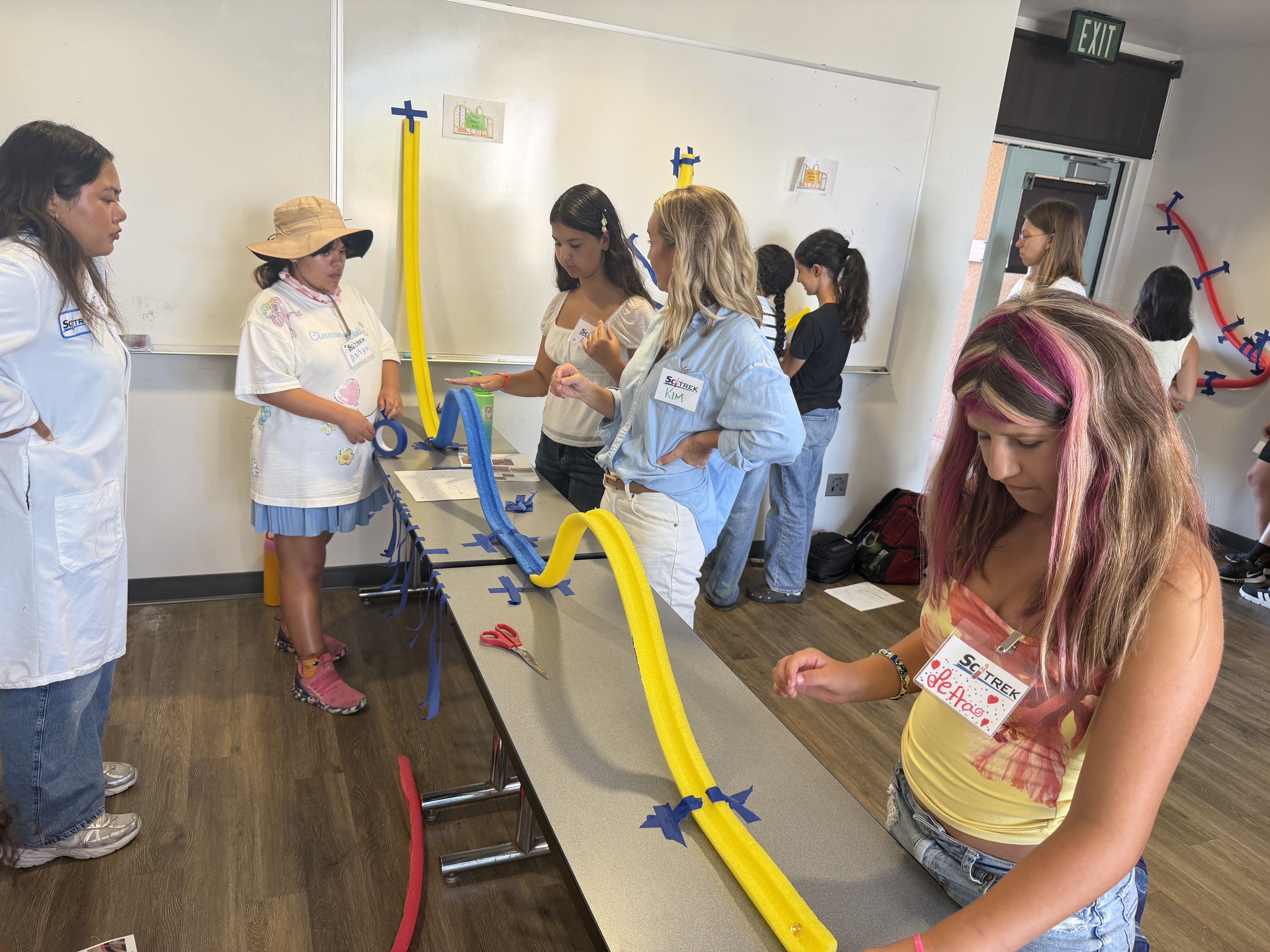 Instructor Kim Ciancio and SciTrek students build a marble roller coaster using blue and yellow pool noodles on a classroom table.