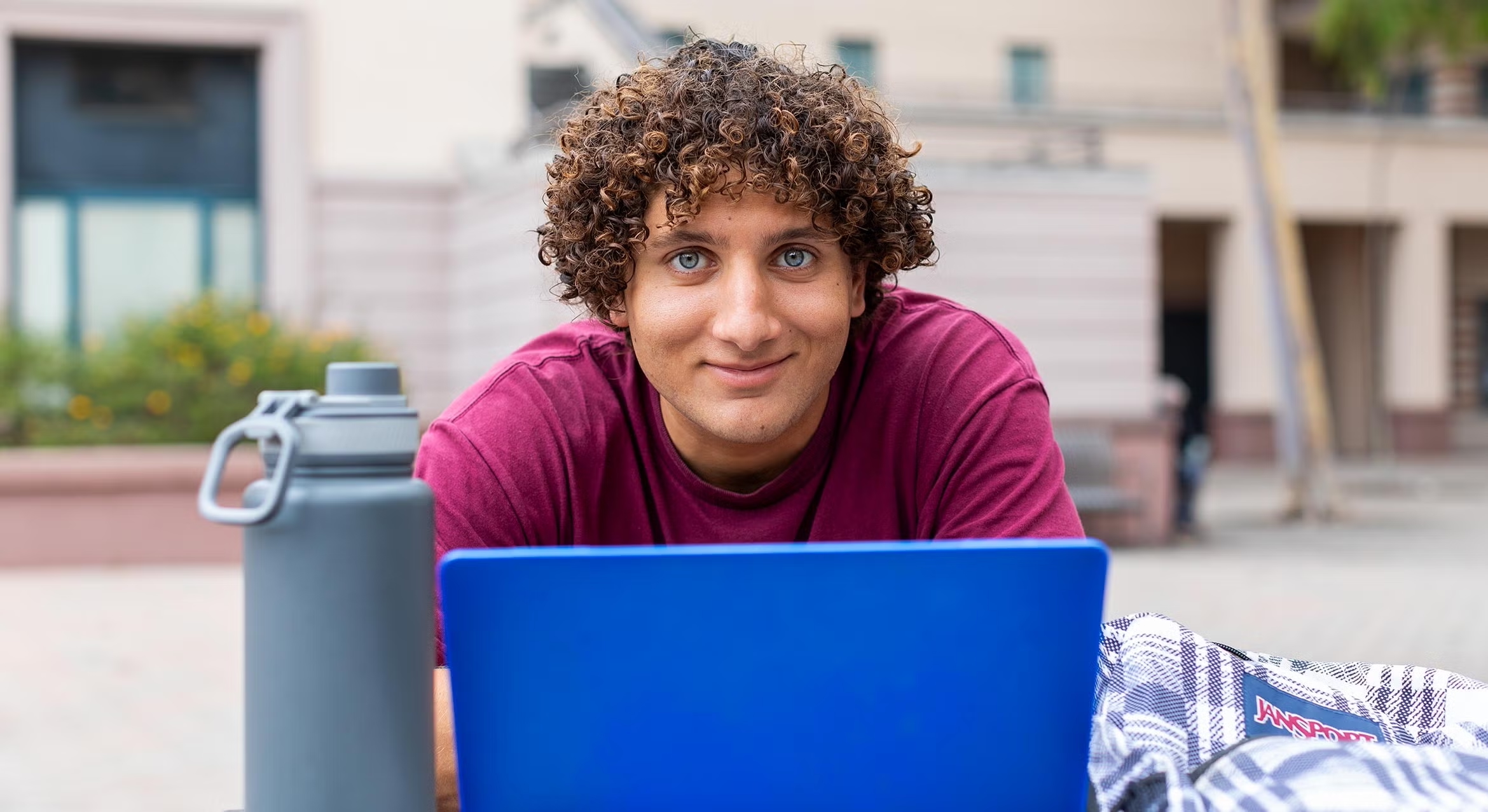A student smiling on campus with a laptop and water bottle.