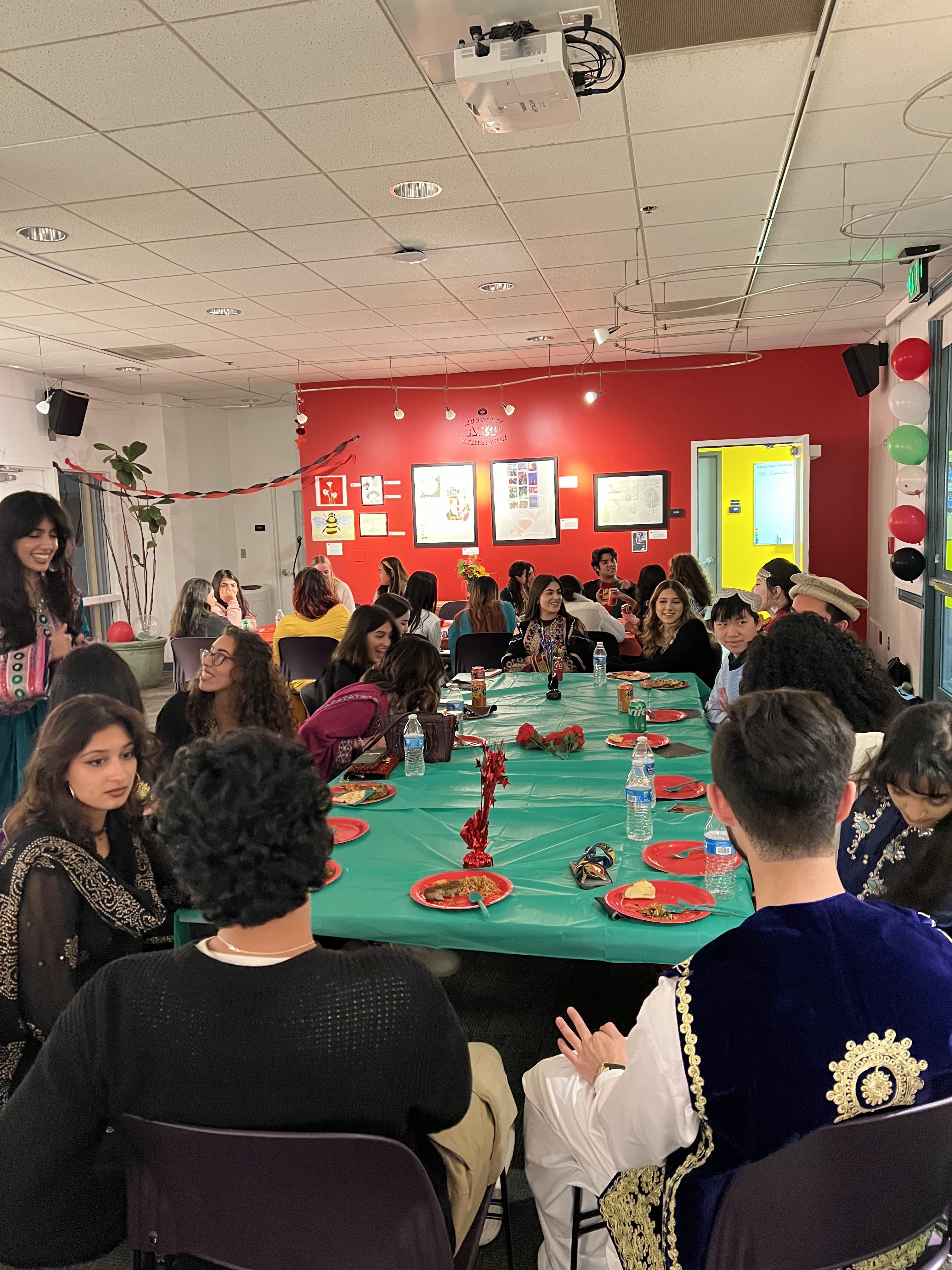 An indoor banquet with students seated around a long green table. Ghazel Durani stands on the left in traditional Afghan dress, smiling and talking with the seated guests. Red, green, and black balloons decorate the background.