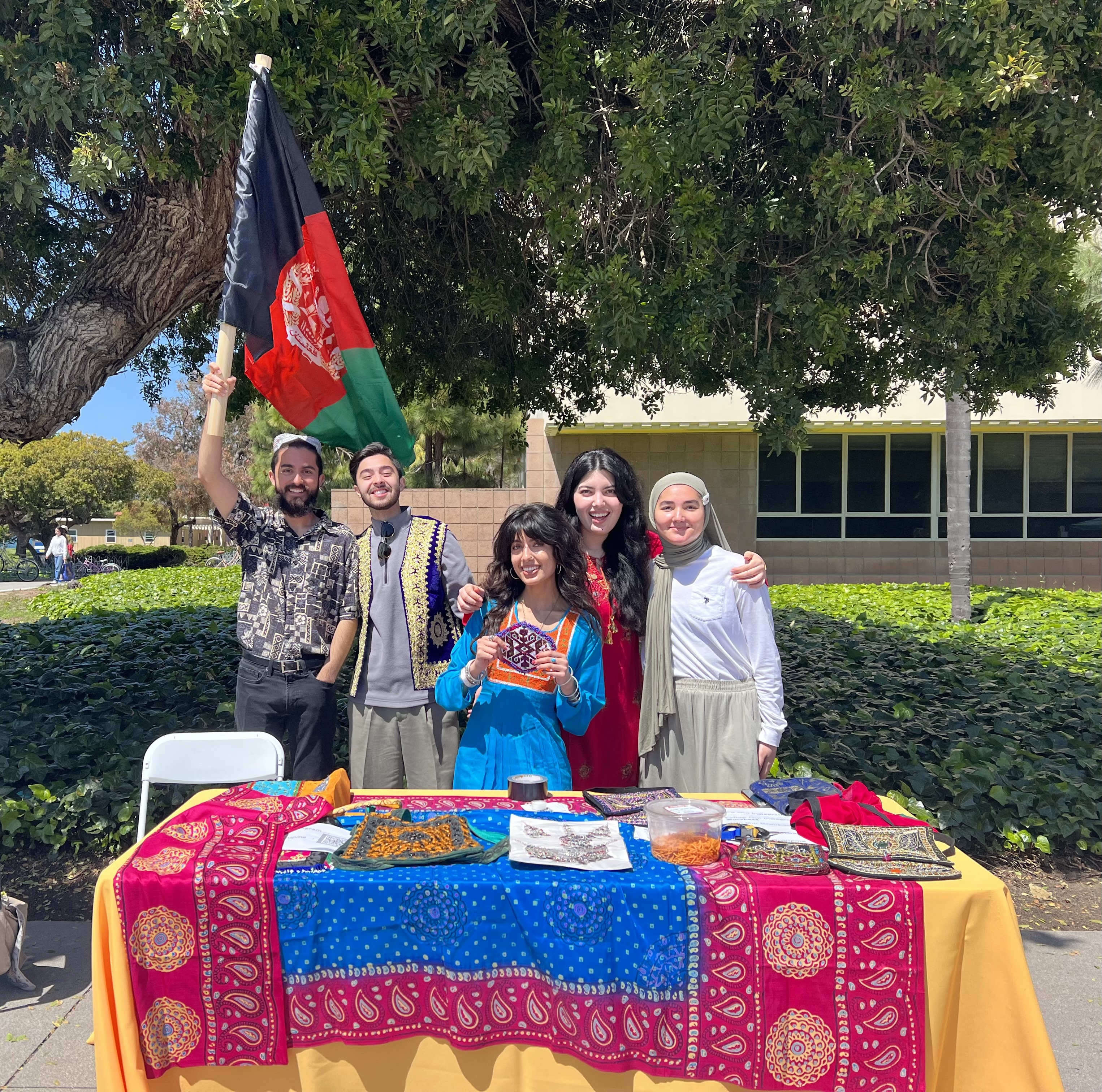 A group of five smiling students standing outdoors behind a yellow table displaying colorful Afghan textiles and cultural items. One student holds up the flag of Afghanistan.