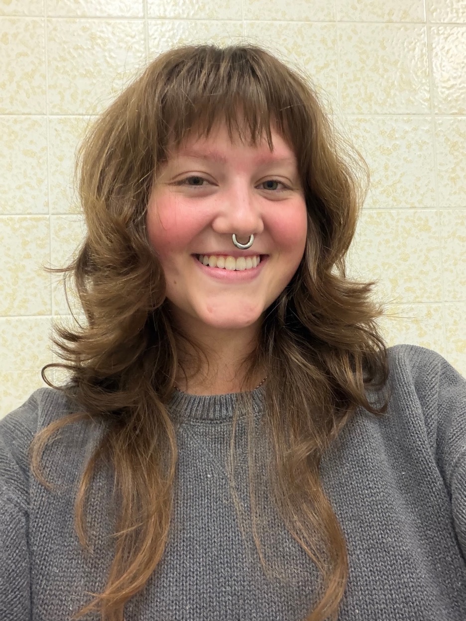 Selfie of Iris Keesey smiling. She has shoulder-length brown hair with bangs and is wearing a gray sweater and a septum piercing.
