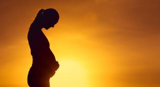 Silhouette of a pregnant woman standing in profile against a warm, golden sunrise, looking down and holding her belly.