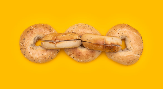 Four bagels arranged in a horizontal line against a bright yellow background. The two middle bagels are stacked, while the outer two lie flat. Each bagel has a light brown crust with poppy seeds sprinkled on top.