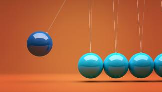 A Newton's Cradle demonstration with five steel spheres suspended in a line, illustrating the principles of conservation of momentum and energy. The leftmost blue sphere is pulled back and held, ready to strike the four stationary light-blue spheres.