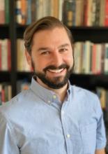 Brendan Bowler, a man with a beard and short brown hair, smiles in a light blue button-down shirt in front of a bookshelf.
