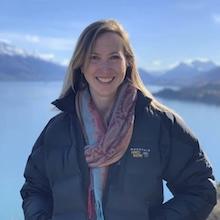 Caitlin Casey, a woman with long blonde hair, smiles in front of a blue lake and mountains in New Zealand.